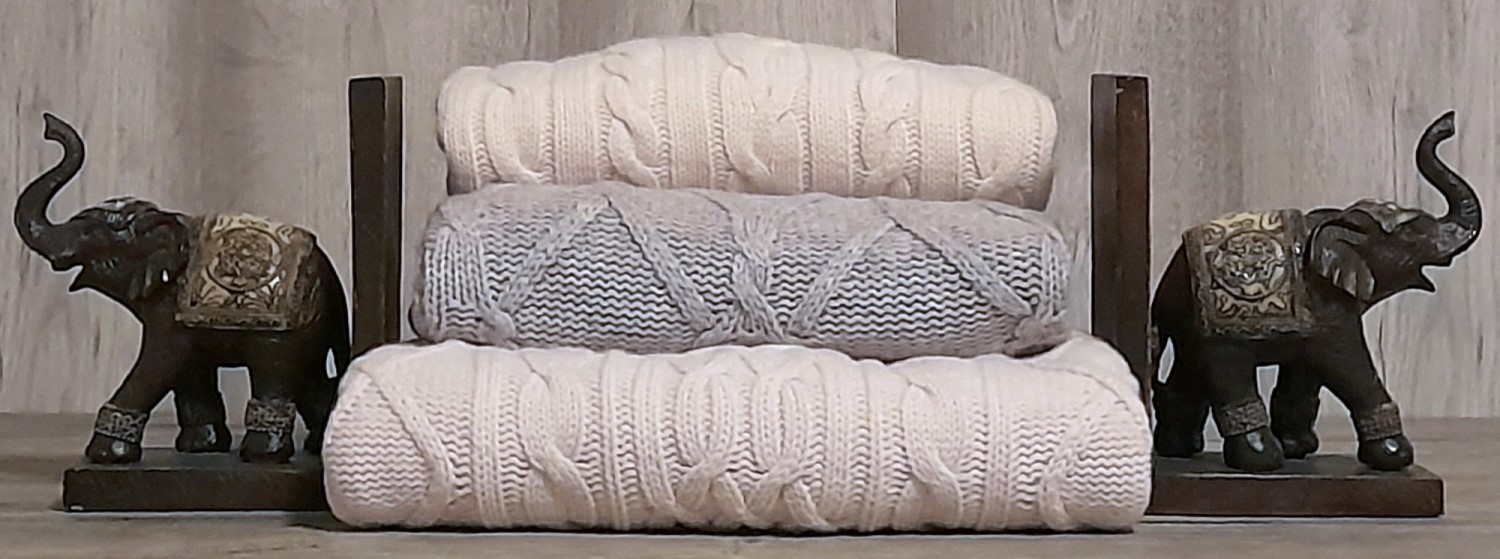 Drei gestapelte Balldiri Cashmere Pullover mit Zopfmuster in Cremeweiß und Grau, stilvoll arrangiert zwischen zwei dekorativen Elefanten-Buchstützen.